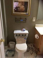 View of toilet area with framed floral print above, metal wall rack mounted above toilet tank, wicker and metal magazine rack on toilet tank, glass dish with bath bombs on toilet tank, ceramic waste basket to the side, and metal curved wall towel rack to the right.