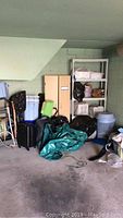 Photo shows corner of a garage with beige storage cabinet, white plastic shelving unit, plastic totes, luggage, and a green tarp on the floor.