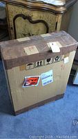 Cardboard box sealed with tape containing the wall cabinet, placed upright on carpet with other furniture visible in background.