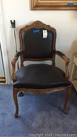 Vintage style arm chair with carved wood frame, dark brown leather-like seat and back upholstery, showing wear on edges and leaf details