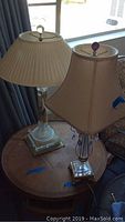 Two lamps on round wooden table near window: clear glass pleated shade and beige shade with purple drops