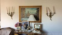 Wide view showing framed painting, table lamp on table, two brass wall sconces with candles on wall, floral arrangement, ceramic dog figurine, and fairy figurine.
