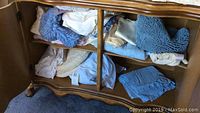View of upper cabinet compartment showing folded assorted linens including light blue microfiber duster, white linens with lace trim, and beige towels.