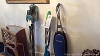 Three vacuum cleaners standing against a wall: an Oreck Magnesium upright, a Shark Rocket stick vacuum, and a Bissell Featherweight stick vacuum.