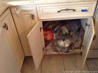 Kitchen cabinet bottom shelf and drawer open showing various pots, pans, and kitchen tools wrapped in plastic bags. Box of Oglettes egg rings visible.