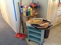 Two wooden furniture pieces against a wall, a shabby chic side table with multiple cleaning supplies on top, a light blue painted rolling cart with trays and stacks of items on it, a mop and broom leaning nearby.