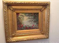 Oil painting with ornate gold frame showing water and red flowers by a nature scene.