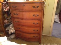 Full front view of wooden chest of drawers showing 5 drawers with curved front and metal handles, in room setting with visible wear.