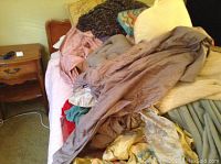 Photo showing assortment of linens and clothing piled on bed with pillows and blankets in neutral and pastel tones.