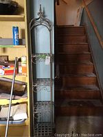 Tall metal rack positioned beside wooden stairs, showing scroll detailing and lattice shelving from top to bottom.