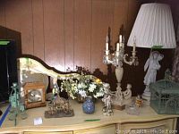 Wide view of mantle-style surface with assorted items: Seth Thomas mantle clock, metal candlesticks, porcelain and ceramic figurines, lamps including cherub prism and porcelain base lamps, wire bandstand decor, faux flowers in metal vase.