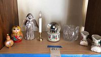 Shelf with glass base vase, silver angel figurine, musical snow globe, candle in gold holder, Russian nesting doll, small wooden gourd container, porcelain shoes with floral designs.
