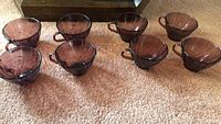 Eight cranberry glass cups with handles arranged on carpet, showing ribbed swirl pattern and clear cranberry color.