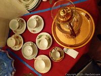 Seven tea cup and saucer sets of various designs placed on a red surface with some floral patterns and gold trims.