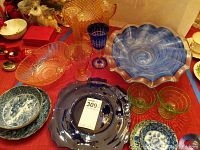 Photo showing a large blue ruffled carnival glass fruit bowl, cobalt blue ruffled platter, pink and blue goblets, amber glass bowl, and oriental style blue ceramic bowls on a red tablecloth.