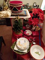 Overall display of holiday decor and dinnerware including drum box, poinsettia, tree, plates, bowls, mugs, and other decorations arranged on red tablecloth.