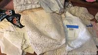 Photo showing a pile of vintage linens including various lace and embroidered pieces, mostly in neutral tones. Includes tablecloths and decorative cloths with intricate lace patterns.