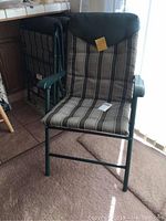 Front view of one folding outdoor chair with striped brown, beige, and cream cushions and green metal frame with plastic armrests. Other chairs folded and stacked behind.