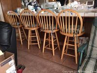 Four wooden swivel bar chairs with light-colored round cushions, arranged side by side under a counter. Cushions show some stains. Chairs have spindle backs and swivel seats.