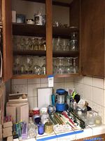 Photo showing wooden kitchen cabinets open with shelves containing multiple rows of assorted wine, beer, and liqueur glasses. Below the cabinet, various kitchen items, candleholders, and candles are visible on the counter.
