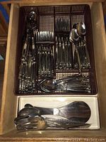 Top view of the stainless steel flatware in a brown plastic tray inside a wooden drawer, showing knives, forks, spoons, and serving utensils.