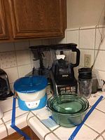 Kitchen countertop with Ninja Auto-IQ NutriNinja blender with two jugs, three green glass mixing bowls nested, and a white and blue Homedics paraffin heater.