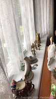 Photo showing brass giraffe figurines, frogs, cats, carousel horse bell, and pitcher on window ledge near sheer white curtains.