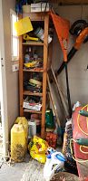 Wide view showing shelving unit with gardening tools including a yellow shovel, orange rake, black and orange leaf blower, plastic containers, gardening bags, and other accessories.
