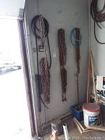 Wide view showing multiple heavy-duty metal chains of various lengths and sizes hanging on wall, including black and blue tubing hung nearby. Floor contains some items like bucket and wooden sticks.