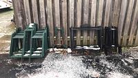 Green and black metal sign frames stacked outdoors on gravel, leaning against a wooden fence, some snow and leaves visible on ground and frames.