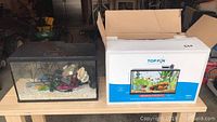 Photo showing one used aquarium with gravel and decorations next to unopened Top Fin Essential 10 gallon aquarium starter kit box.