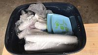 Top-down view of a plastic bin containing stacks of disposable white cups, green disposable plates, and plastic bags filled with other disposable items.