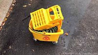 Yellow commercial mop bucket with wringer attachment and black handle, showing green hose coiled inside and four caster wheels on black asphalt ground.