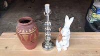 Overall view of all three items on wooden surface: bunny figurine, clay vase, and glass lamp base.