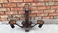 Front view of rustic metal dual candle holder with curled arms and two scalloped edge candle cups, showing rust and aged patina on concrete floor in front of brick wall.