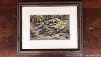 Framed watercolor landscape artwork showing rocky stream and greenery.