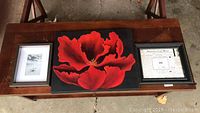 Photo of three framed art items placed on a bench, including a large red poppy print in center, black and white photo on left and framed certificate on right.