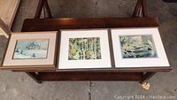 Three framed prints displayed horizontally on a wooden surface, showing outdoor nature and rural scenes.