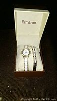 Closed box containing an Armitron watch and two bracelets displayed on a white cushion.
