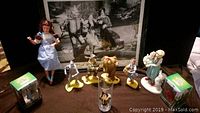 Full display of Wizard of Oz porcelain dolls, character figurines, glass and picture in background.