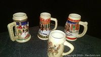 Four ceramic Budweiser holiday steins displayed on a dark surface, showing detailed winter scenes and festive accents.
