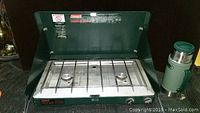 Coleman two burner propane stove with green lid open showing metal burners and grate