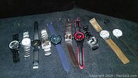 Full lot of 10 men's wristwatches arranged in a row on dark surface, showing variety of bands and face colors.
