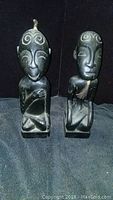 Photo of two dark stone African statues with stylized faces and spiral forehead motifs on a dark fabric background.