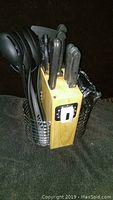 Wooden knife block holding several black-handled knives, black plastic utensils, and a pair of kitchen scissors seen from the front