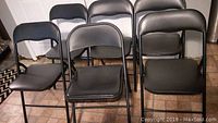 Six black folding chairs arranged standing up against a wall, showing padded seats and backs and metal frames.