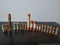 Full view of 16 cylindrical fuses of various sizes and labels arranged in two rows on gray surface