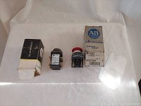 Two Allen Bradley industrial electrical components with original boxes. One component is a cluster pilot light with wiring terminals and product details on the label visible. The other is an emergency stop button with a red top. The black box is labeled Allen-Bradley and is alongside the components. The white box shows the AB brand and model number 800T-D8A.