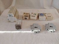 Assorted Allen Bradley electrical components including relays, relay sockets, terminal blocks, some in original boxes, arranged on white cloth background.