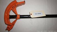 Side view of pipe foot bender showing orange curved bending shoe and black handle with SKU tag.